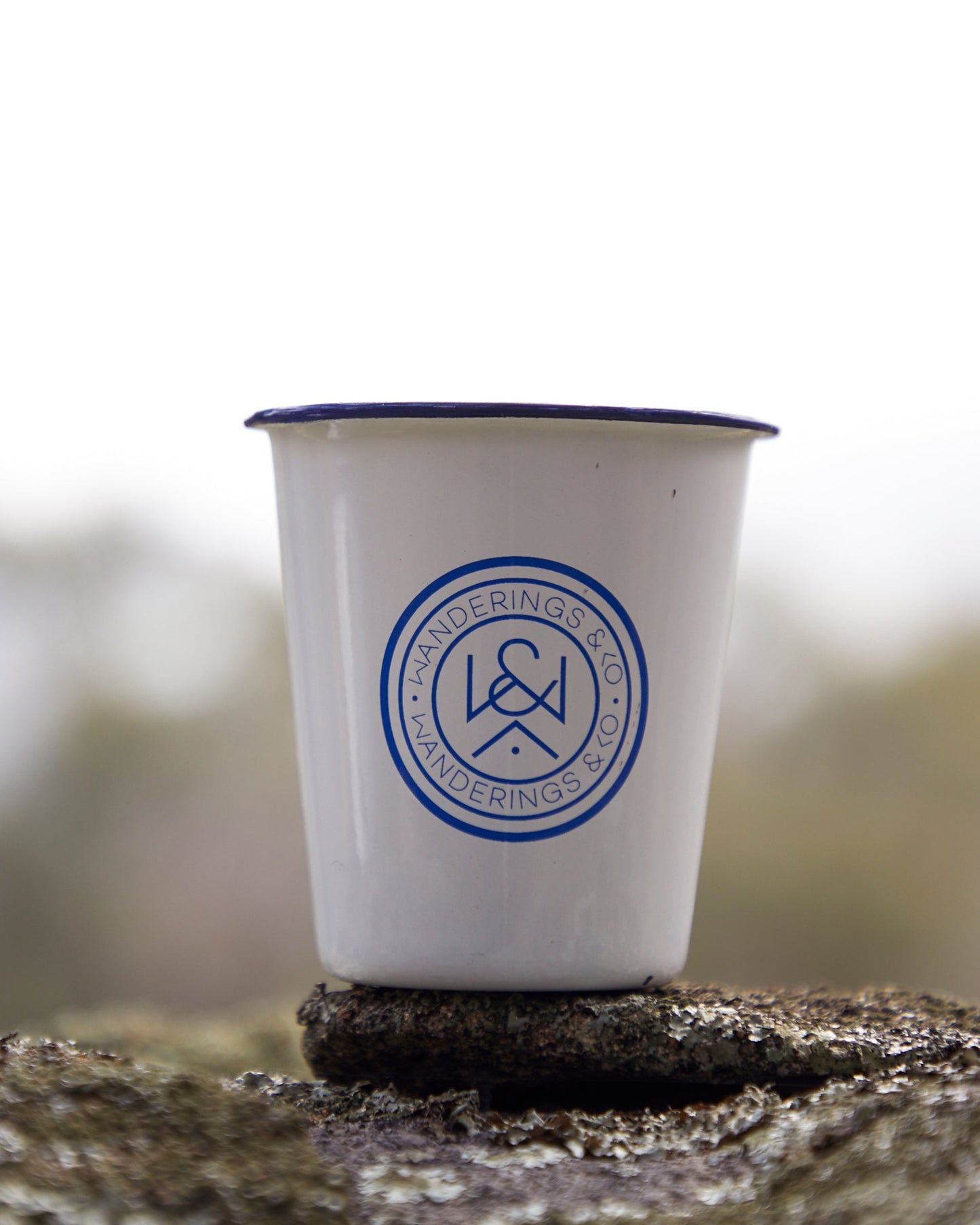 White cup with blue rim and logo on a rock with a blurred natural background