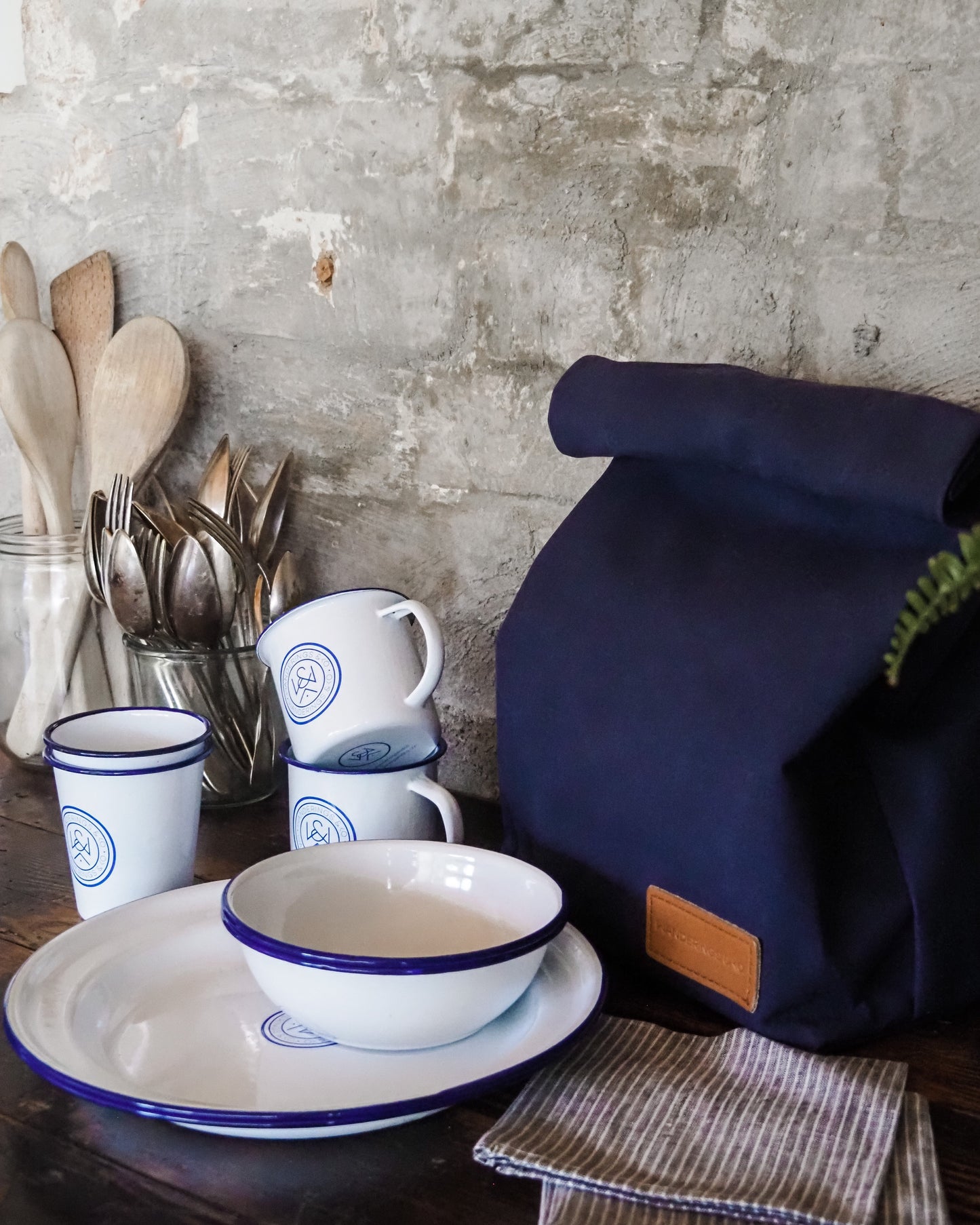 Navy blue backpack on a wooden table with white dishes and utensils against a rustic brick wall.